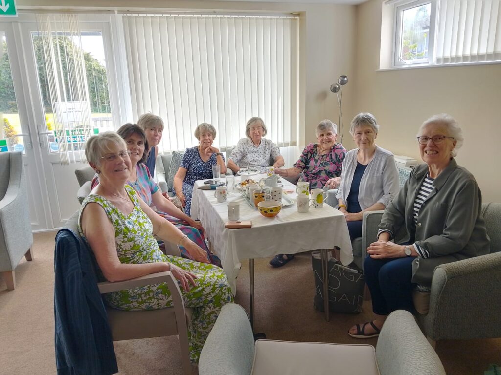 8 happy people sitting around a table that has the leftovers of afternoon tea.